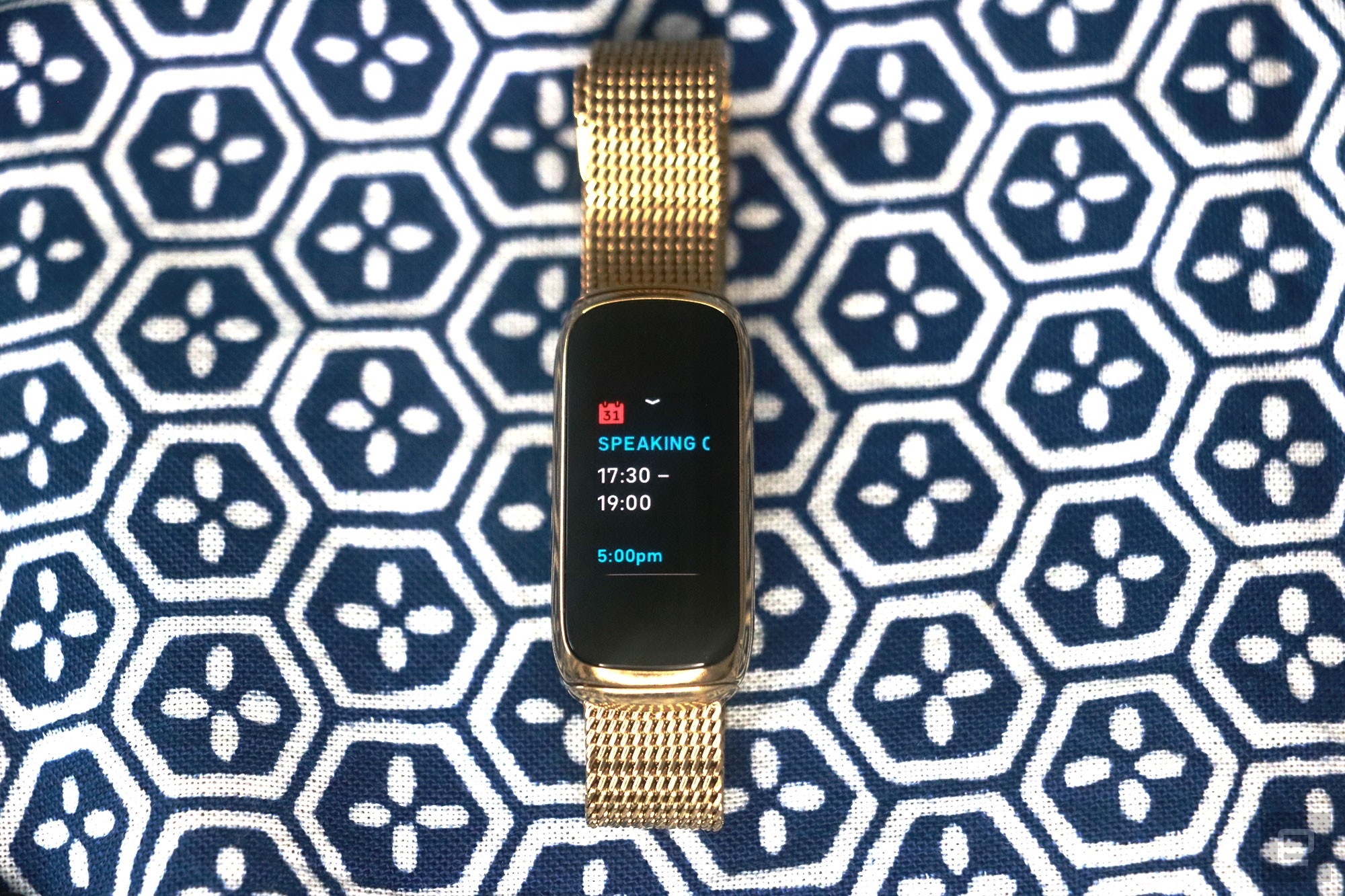 Front view of the Fitbit Luxe with a gold mesh bracelet on a patterned blue and white background. Its screen shows a calendar notification for an event from 5:30pm to 7pm. | DeviceDaily.com