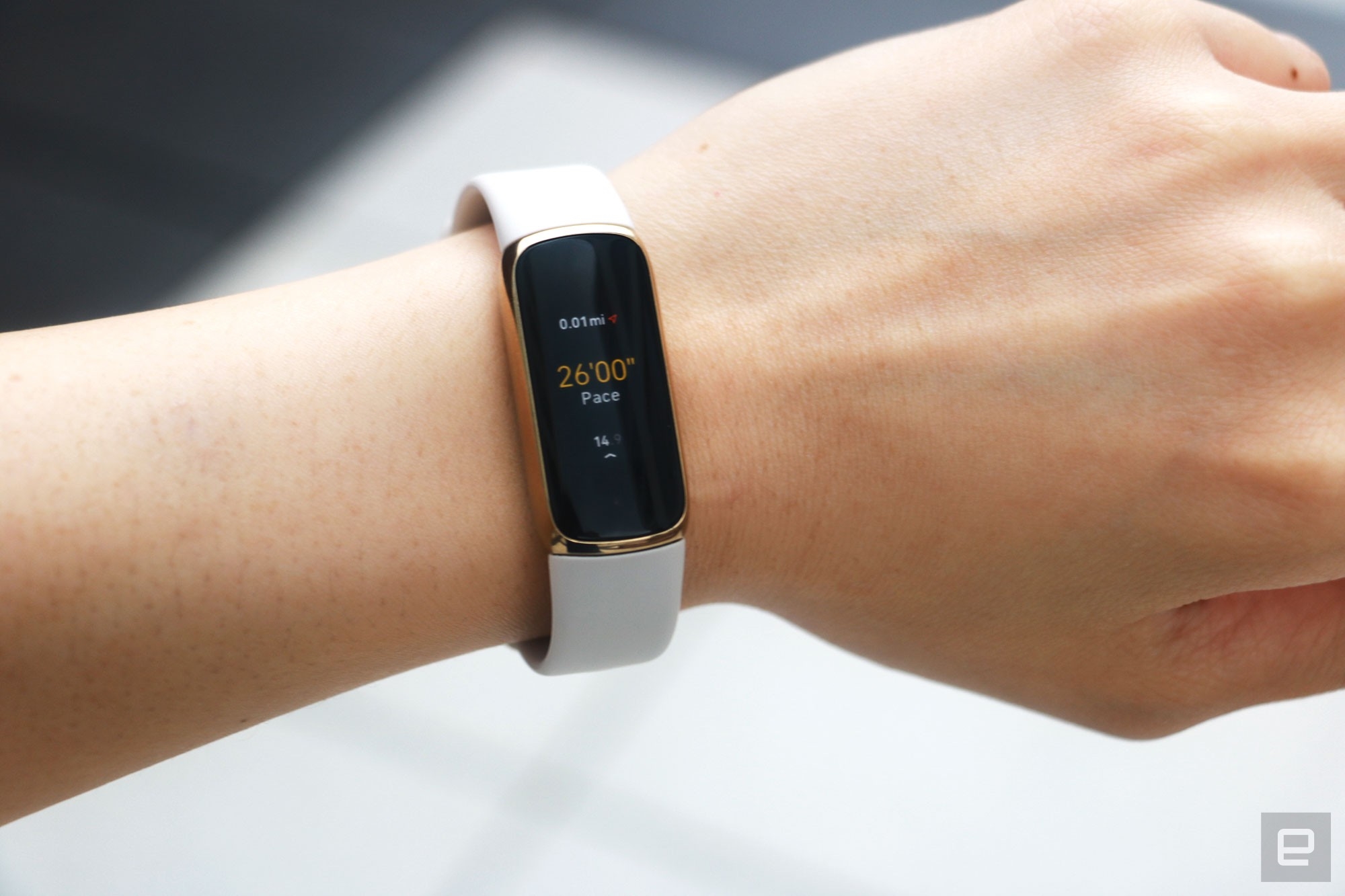 The Fitbit Luxe with a light pink silicone band on a wrist against a concrete gray background. The screen shows a run being tracked with a pace of 26:00 and 0.01 miles traveled. | DeviceDaily.com