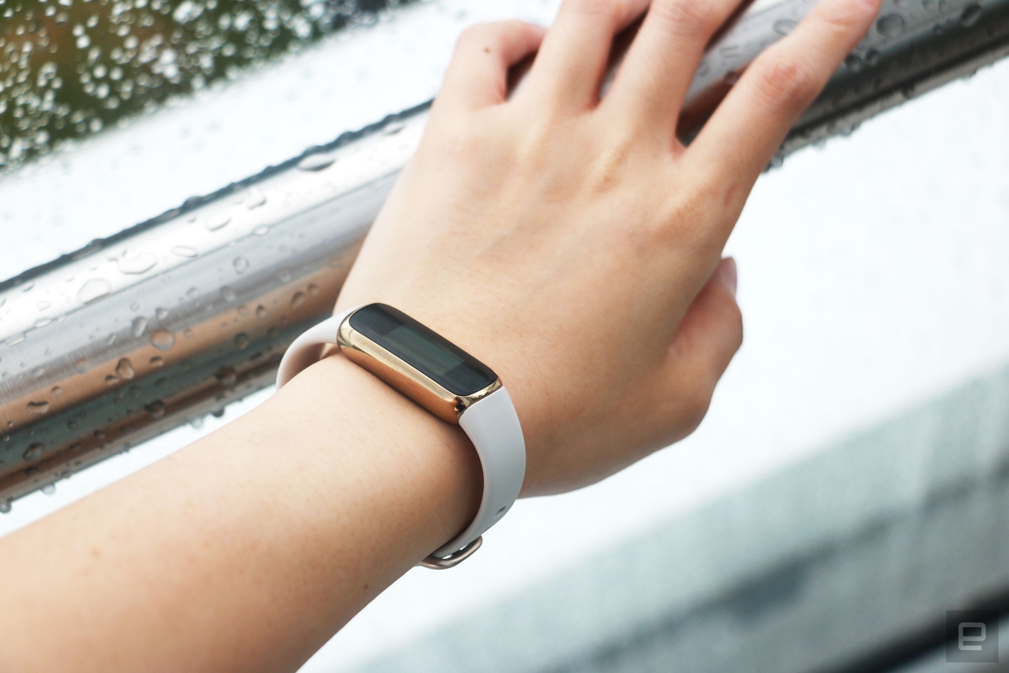 The Fitbit Luxe with a light pink silicone band on a wrist leaning on a wet railing. The screen is off. | DeviceDaily.com