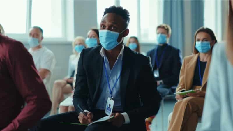 A man sits in the crowd, wearing a surgical mask, at an in-person event. | DeviceDaily.com