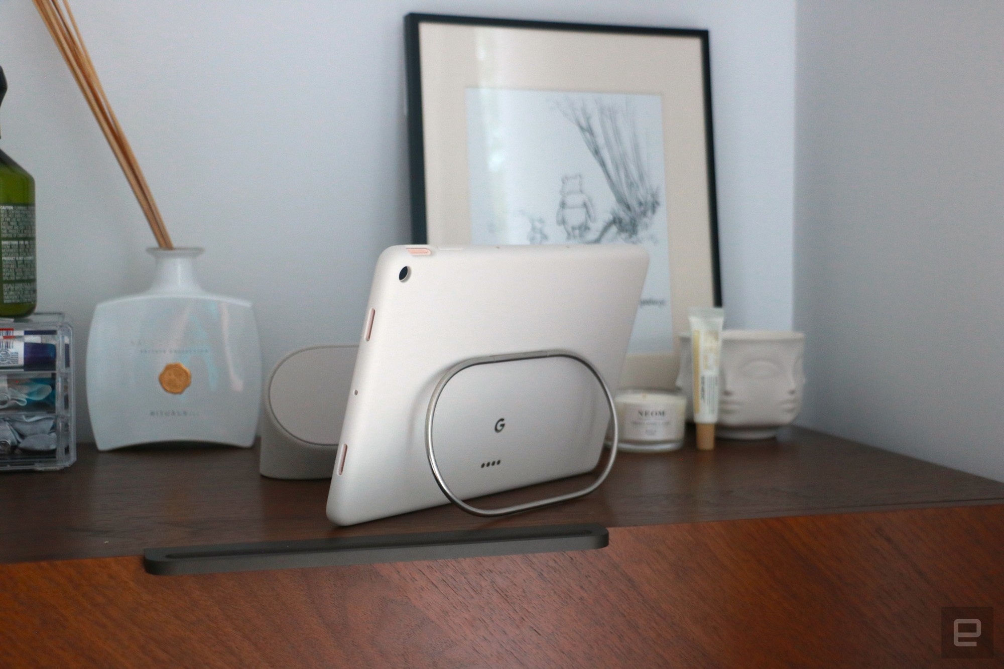 The Pixel Tablet on a walnut dresser propped up by the silver kickstand on the back of its protective case. | DeviceDaily.com