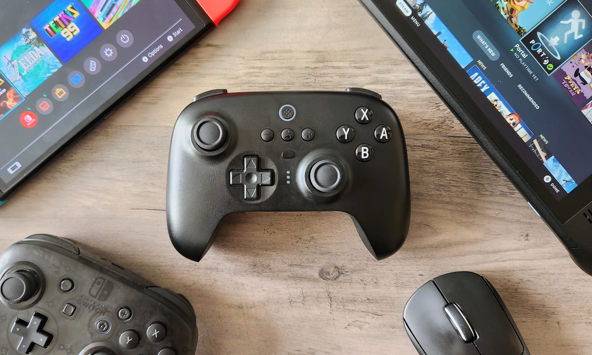 A black video game controller, 8BitDo Ultimate Bluetooth Controller, rests on a wooden table alongside a Nintendo Switch, a Nintendo Switch Pro Controller, a Valve Steam Deck, and a Logitech gaming mouse. | DeviceDaily.com