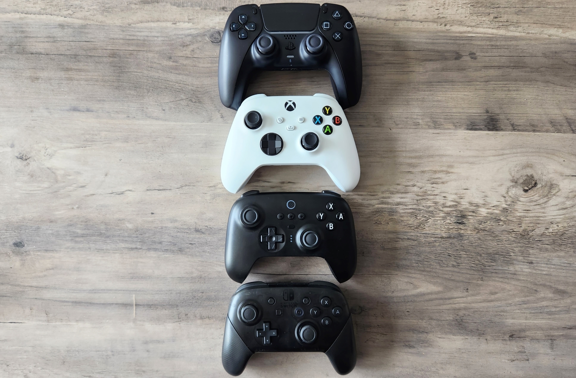Four video game controllers rest on a wooden tabletop, one above the other. From the top: the Sony DualSense PlayStation 5 Controller, the Microsoft Xbox Series X/S Controller, the 8BitDo Ultimate Bluetooth Controller and the Nintendo Switch Pro Controller. | DeviceDaily.com