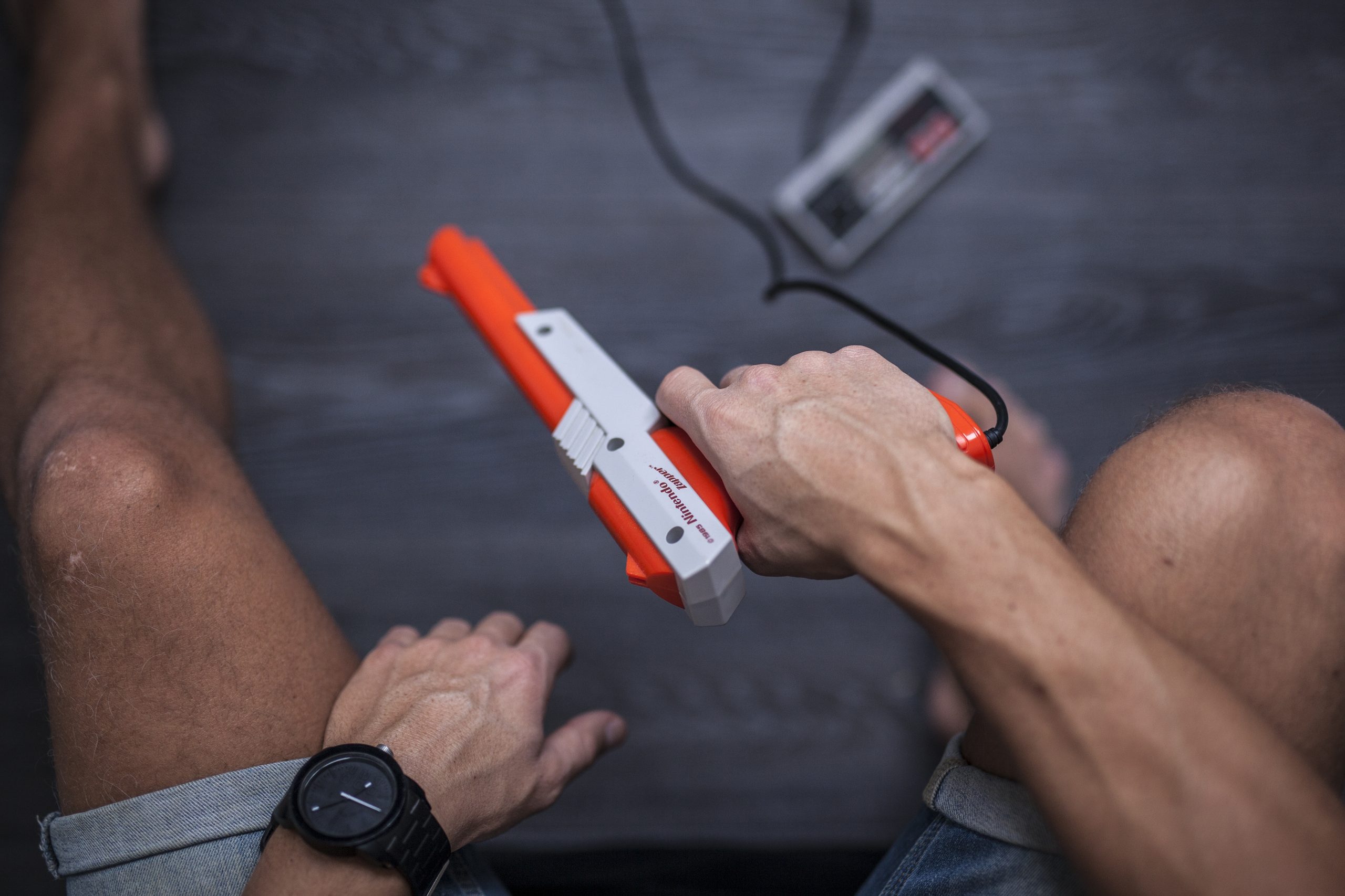 Gothenburg, Sweden - January 31, 2015: A shot from above of a young man's hands using a 1985 Nintendo Zapper, a remote game controller for the Nintendo Entertainment System developed by Nintendo Co., Ltd. in the 1980s. His hands are posing casually as he is resting between games. Natural lighting. Shot on a grey wooden background. | DeviceDaily.com