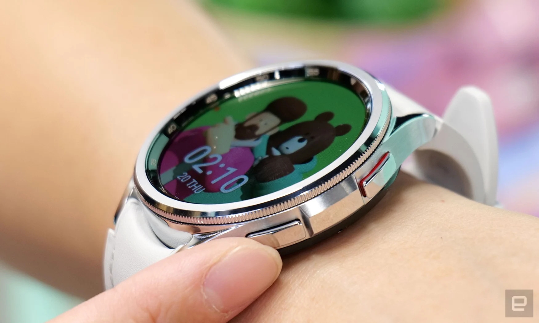 Closeup photo of a Samsung Galaxy Watch 6 smartwatch on a person's wrist. A finger is touching one of the hardware buttons as an image of a man with a teddy bear displays on its screen. | DeviceDaily.com