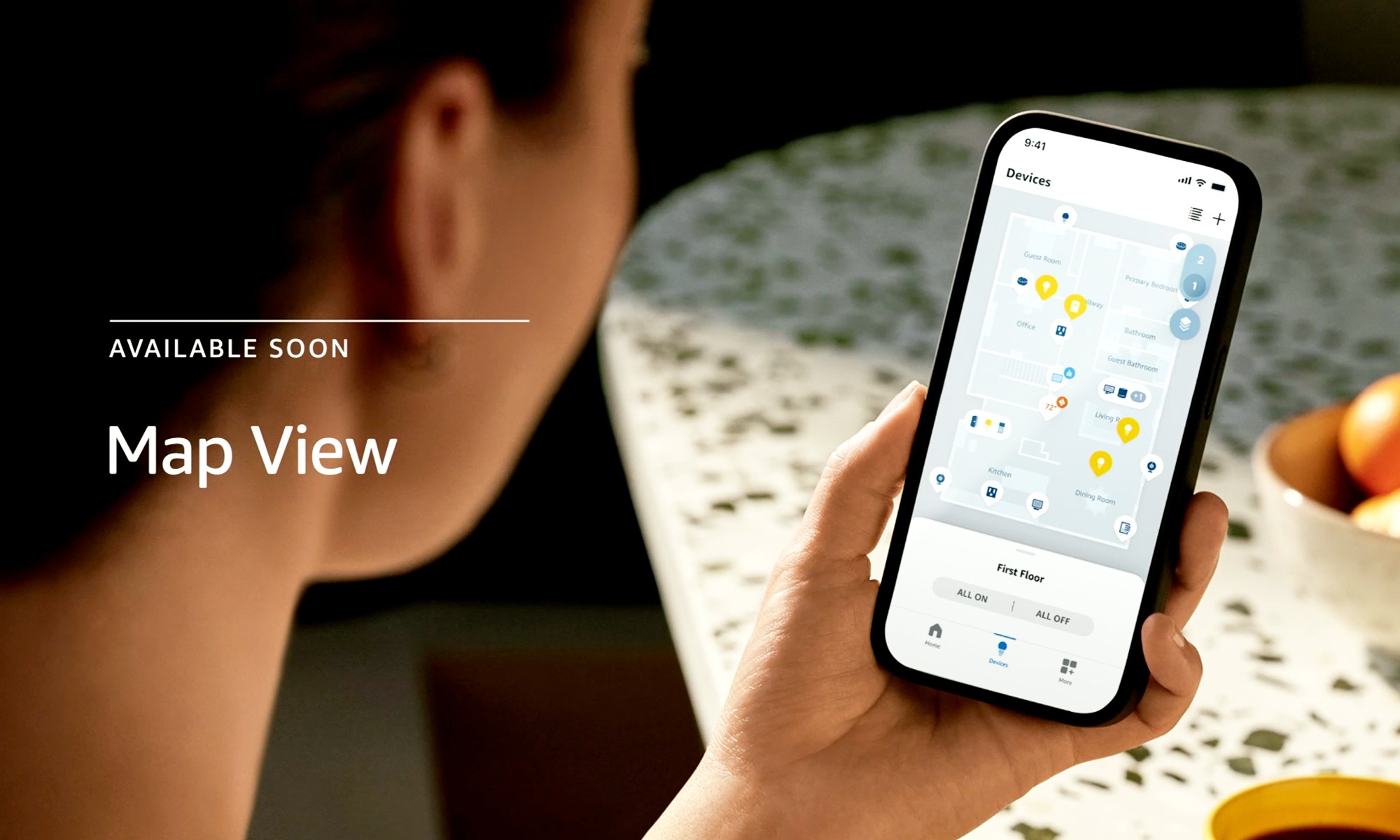 Amazon product photo showcasing Map View. View from over a person's right shoulder as they hold a smartphone. The phone's screen shows a digital floor plan with icons representing smart devices around it. | DeviceDaily.com