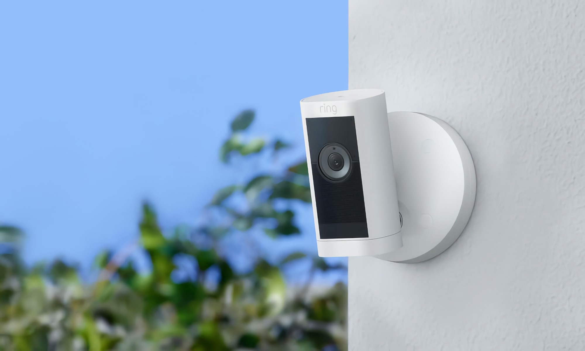 Amazon product photo of the Ring Stick Up Cam Pro. The security camera has a white body and black area near its lens. It's mounted on an outdoor stucco wall with blue sky and green trees visible on the blurred background. | DeviceDaily.com