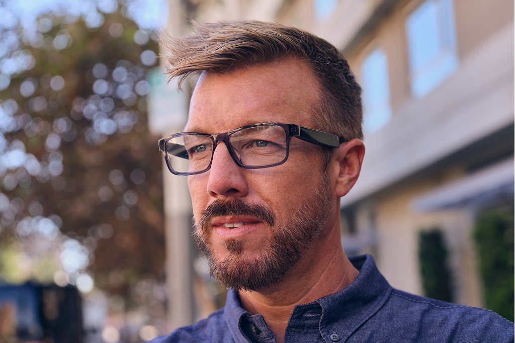 Amazon product lifestyle photography of a man wearing Amazon Echo Frames. The black frames look mostly like regular prescription glasses but have very thick arms. He is middle-aged and has a beard and stylish hair. Outdoors with a blurred background. | DeviceDaily.com